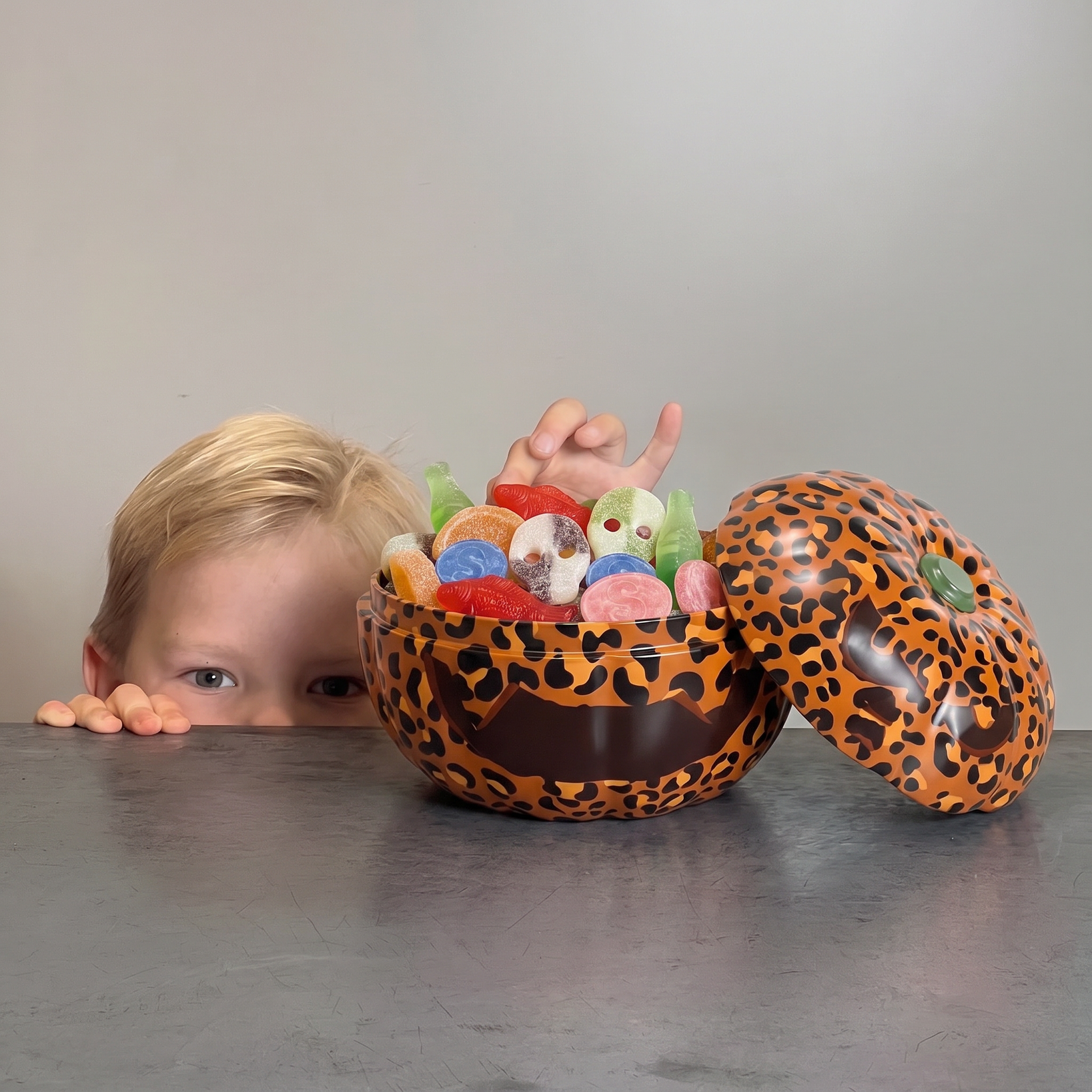 A child with blonde hair peeks over a gray table at a decorative pumpkin-shaped tin with the lid off revealing it is filled with colorful swedish candies. The tin has an orange and black leopard print pattern with a spooky halloween face